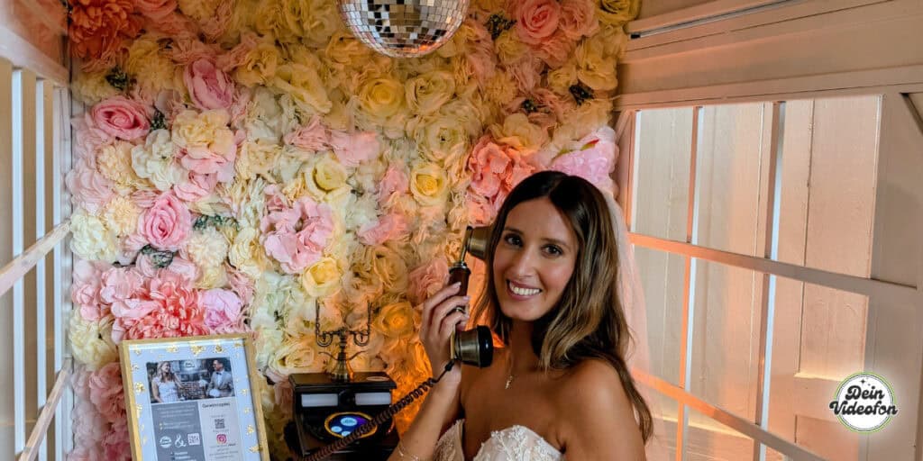 Braut telefoniert vor Blumenwand in Telefonzelle mit einem Videofon. Sie hinterlässt Grüße in das vielleicht modernste Gästebuch der Welt