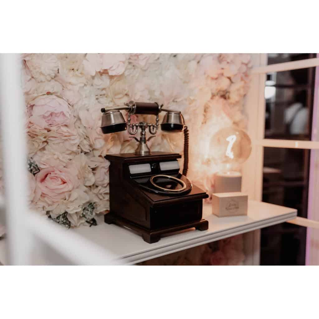 Ein altes Drehtelefon steht auf einem Regal neben einer leuchtenden Glühbirne, vor einem floralen Hintergrund mit großen weißen und rosa Blumen.